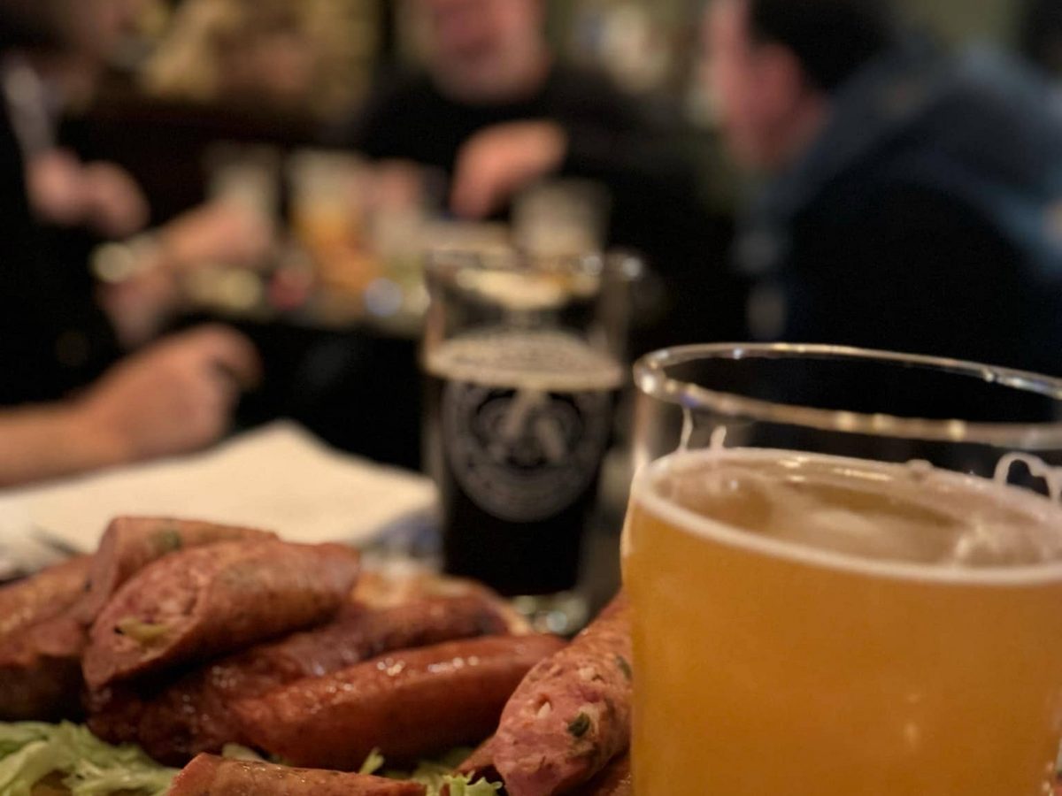 A glass of cloudy beer and a board of grilled sausages on the table at the brewery
