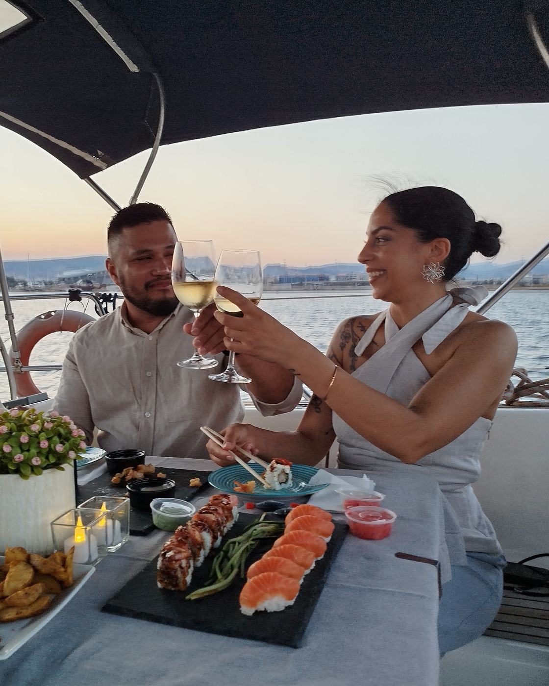 A couple touching glasses of white wine over a table of sushi in the cockpit, the sun going down behind them