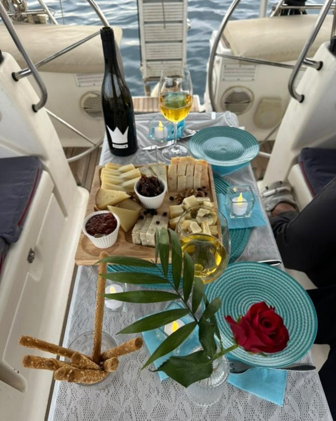 A table set for two on deck: a board of cheeses, an open bottle, a candle and a red rose on the plate