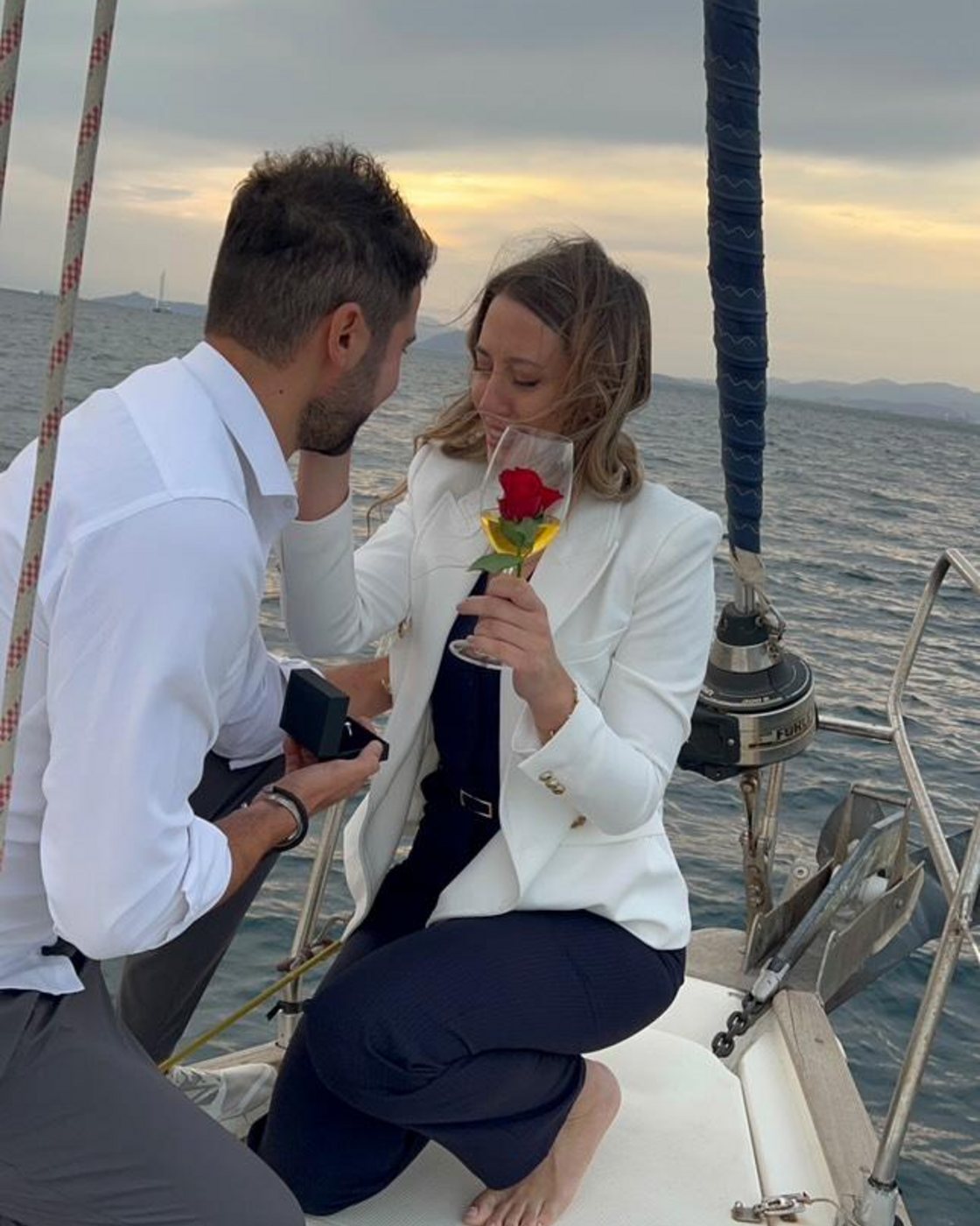 On the foredeck at dusk: he is down on one knee with the box, she is holding the rose from the table