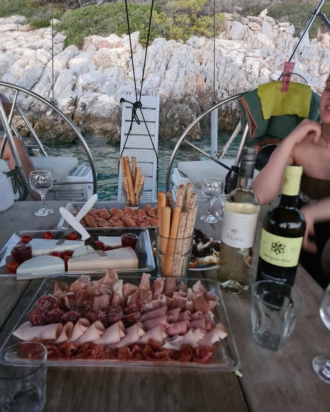 Platters of cold cuts and cheese on the cockpit table at anchor, breadsticks and two open bottles of white, the rocks of the cove behind