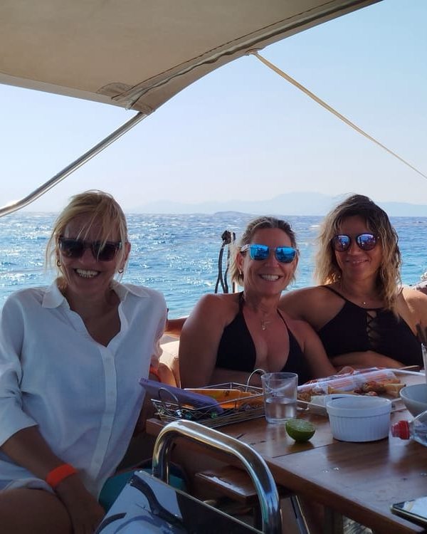 Three guests at the cockpit table under the bimini, open sea and an island on the horizon behind them